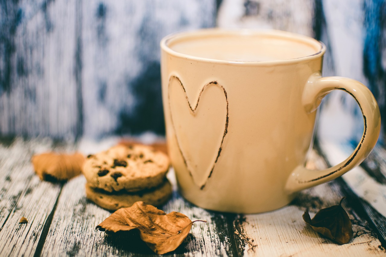 Photo of a mug with a love heart on it filled with coffee next to some leaves and cookies on a wooden backdrop